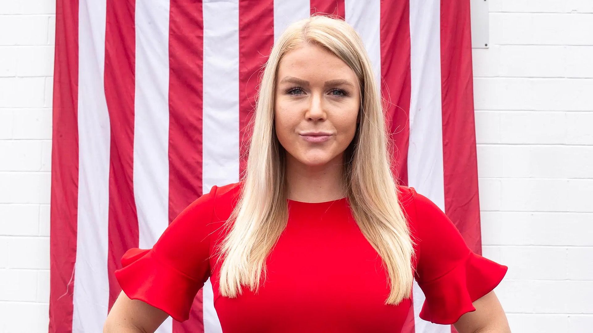 A long-haired blonde in a red dress stands against the backdrop of the American flag