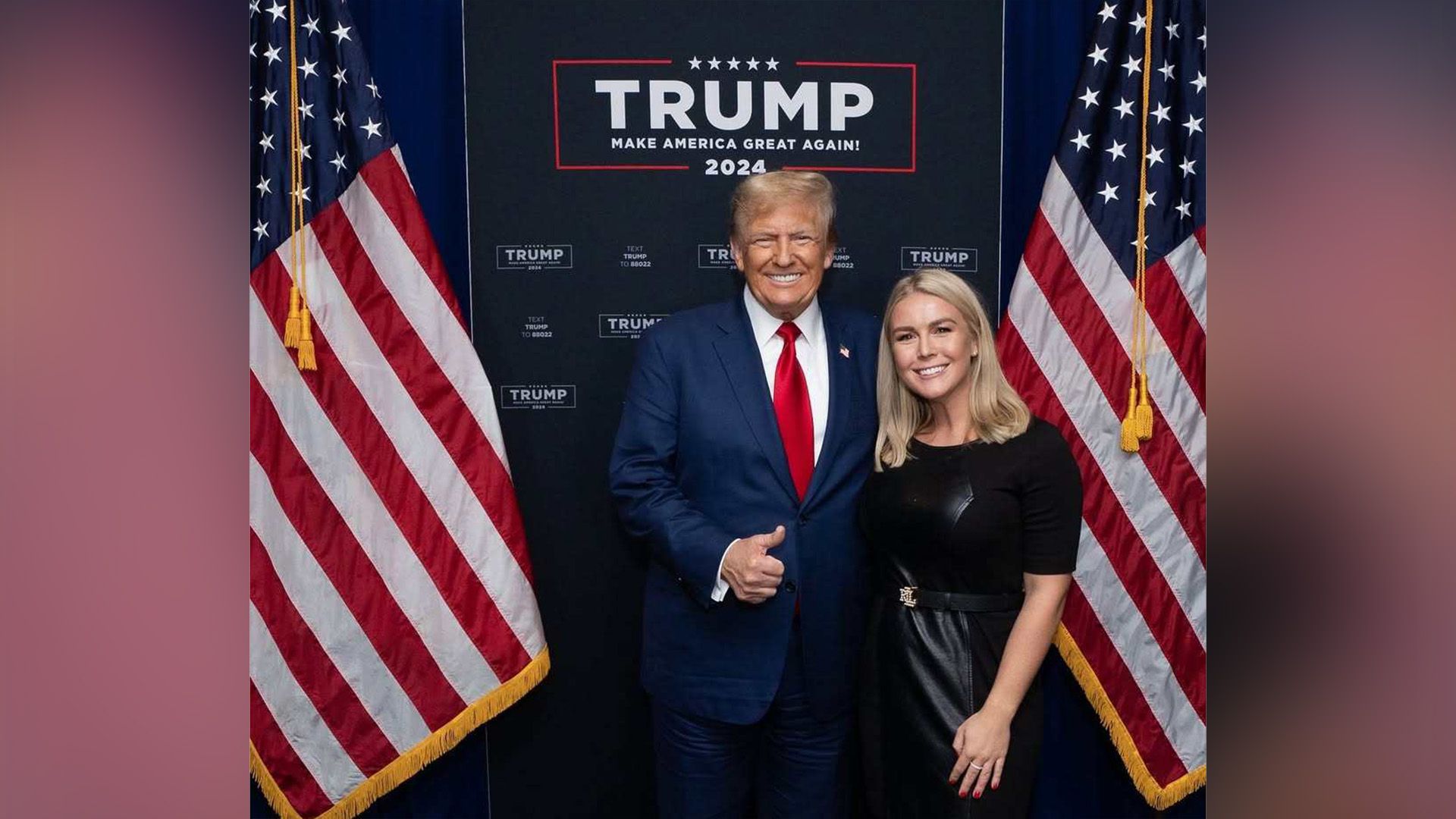 A tall elderly man and a young woman are standing between two American flags, both smiling