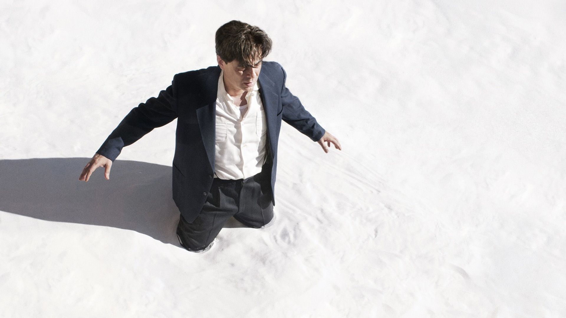 A man in a gray suit is kneeling on white sand, spreading his arms to the sides