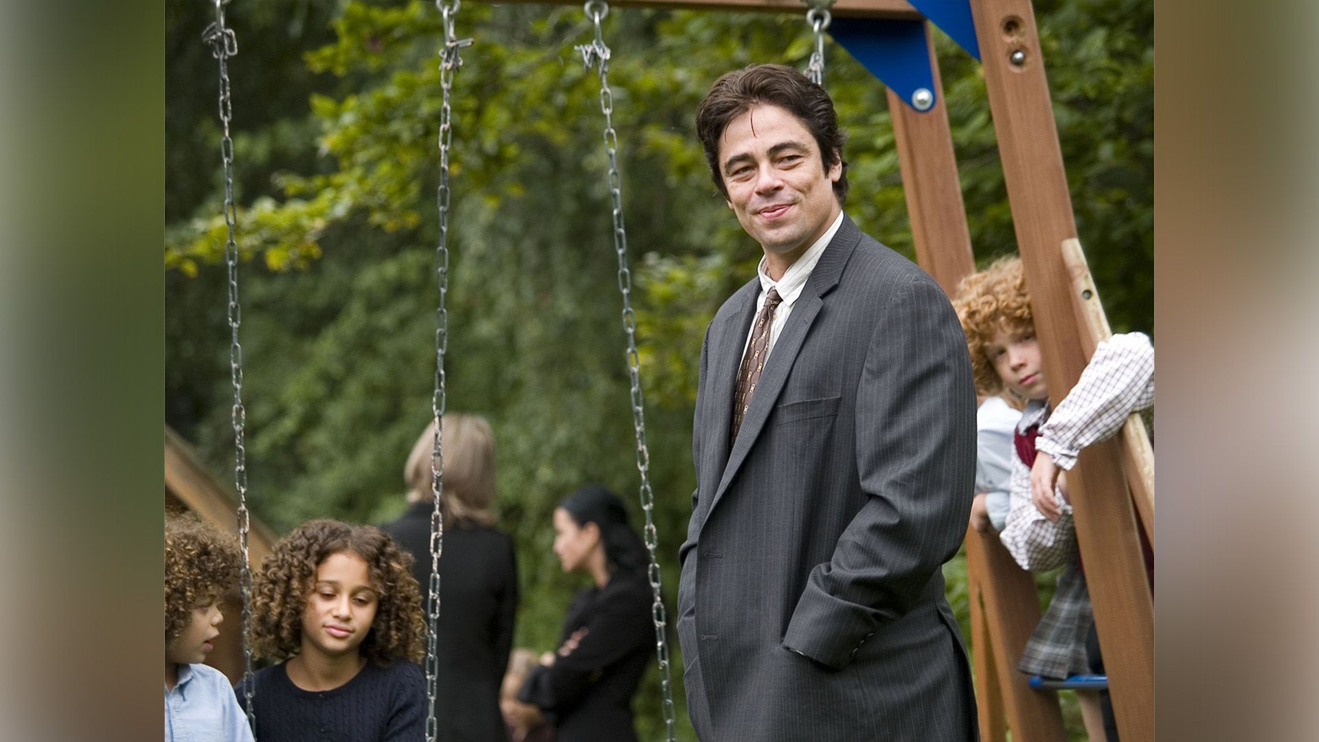 A smiling man in a gray jacket on a playground with several children in the background