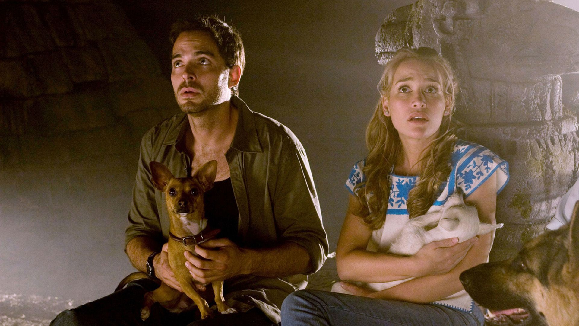 A young man and woman with frightened faces sit in a cave, each holding a small dog