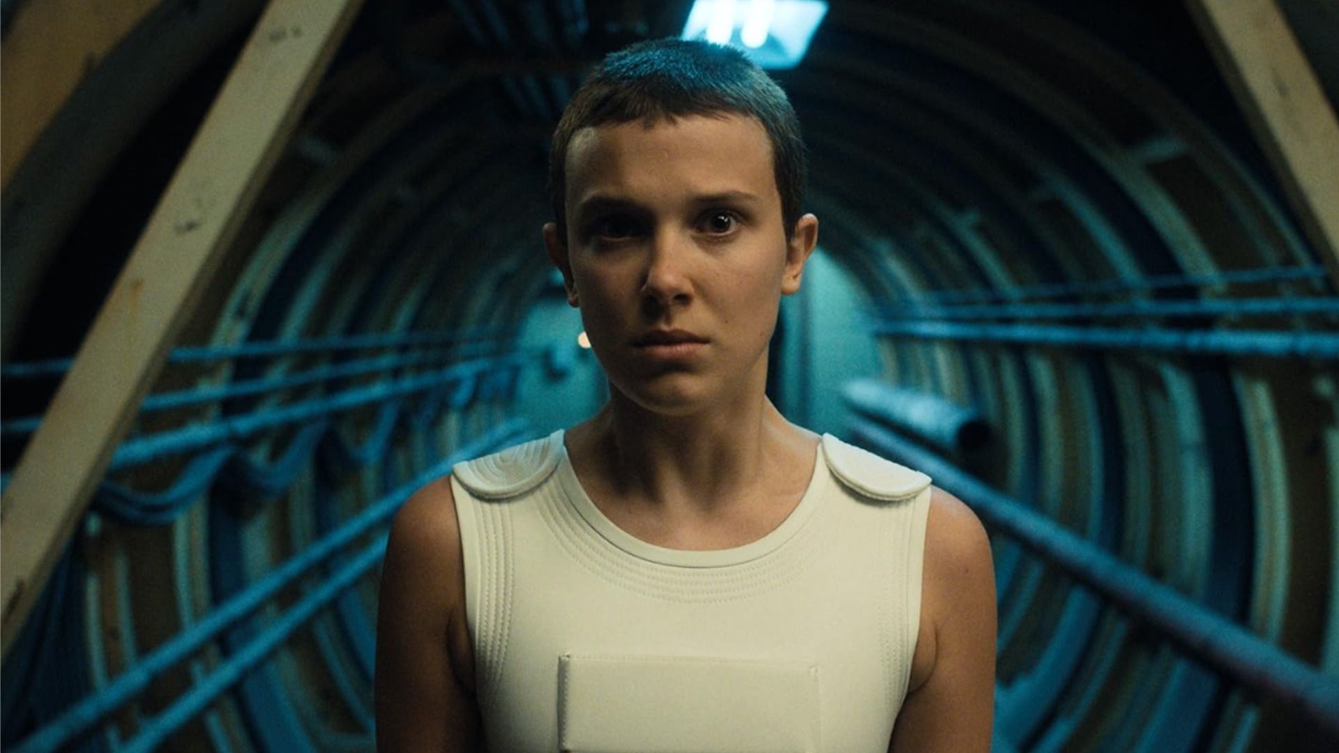 Millie Bobby Brown with a short haircut is standing in a tunnel and looking at the camera