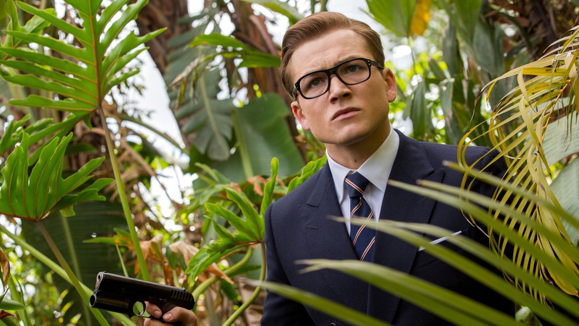 A young man in a jacket and glasses among tropical thickets, holding a pistol in his hand