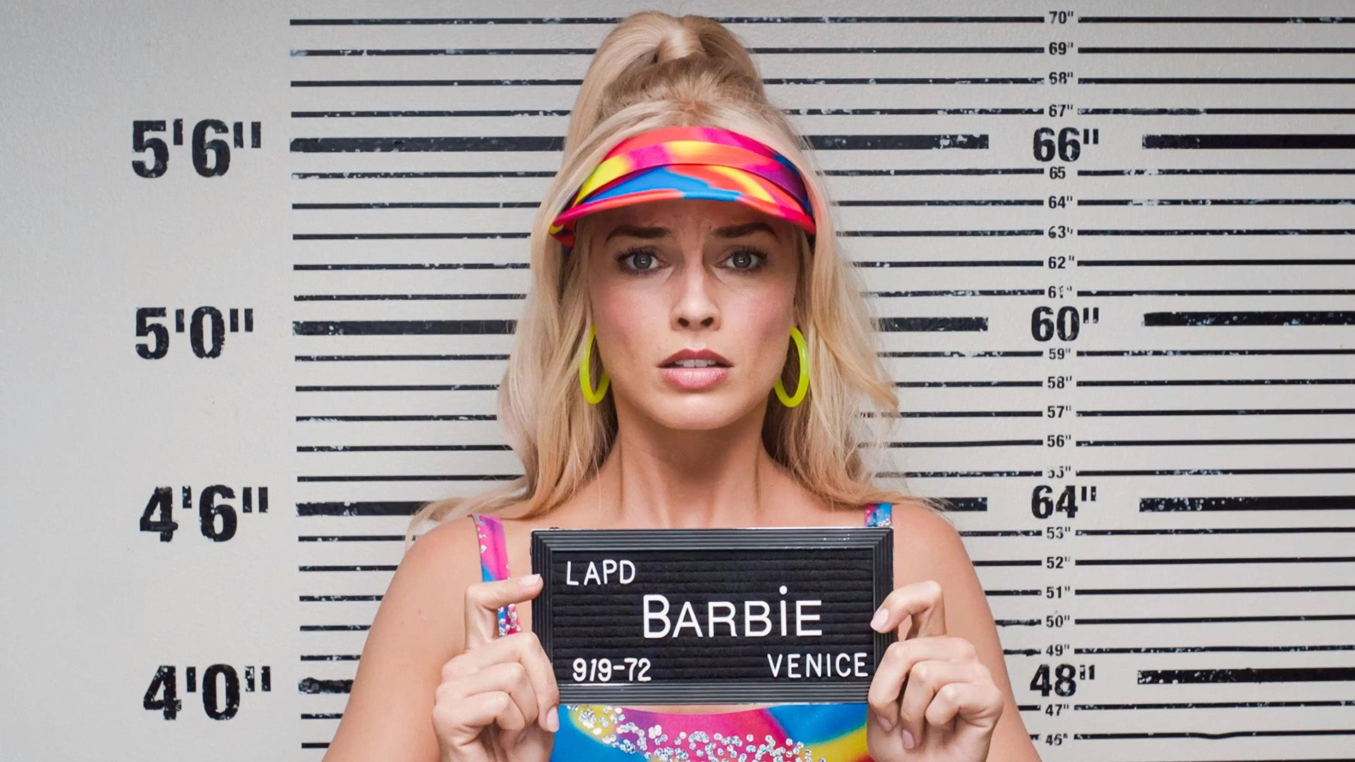 Police photo: a blonde in a bright visor stands against a height chart holding a sign with the name "barbie"