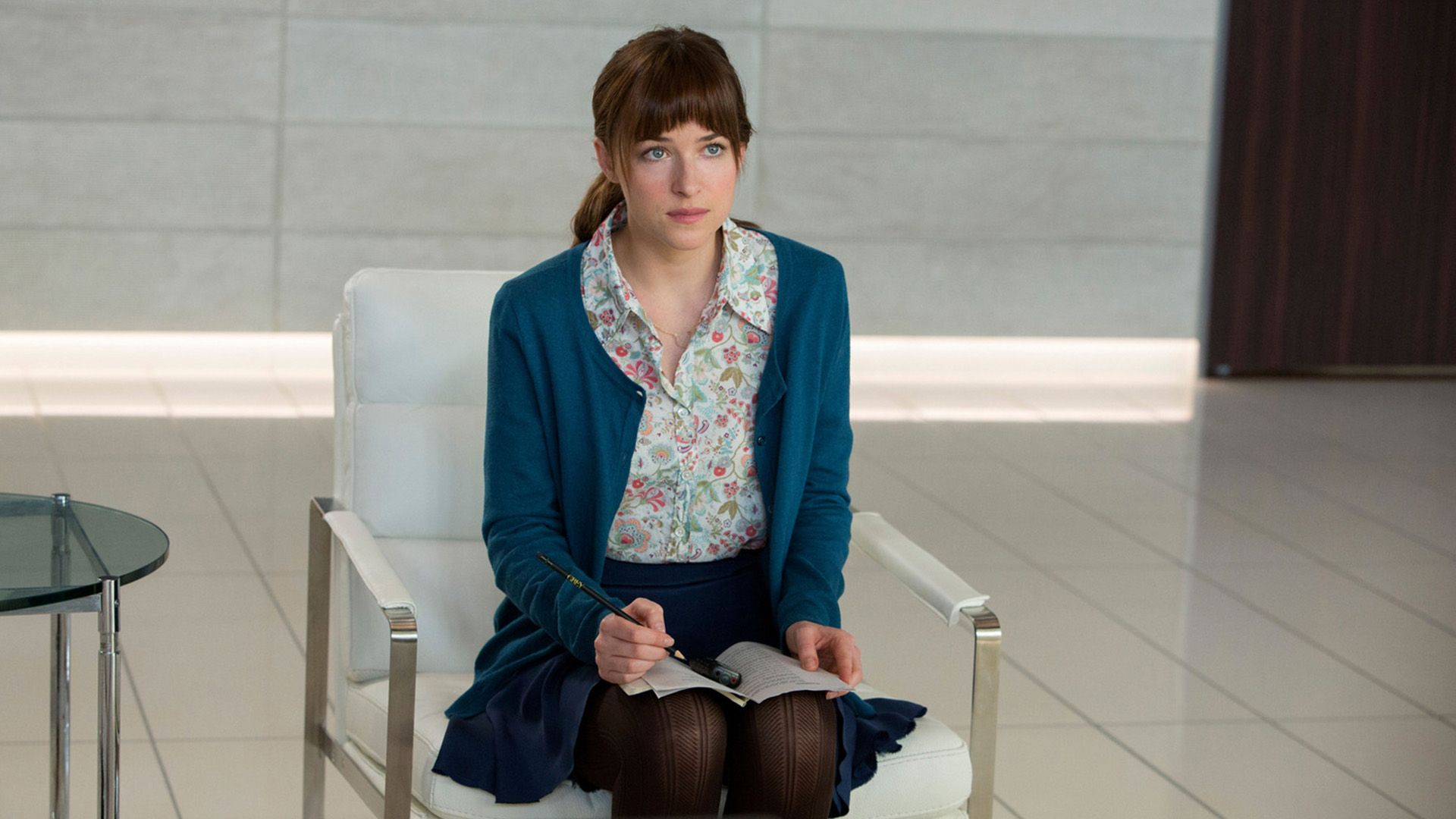 A girl in a blue sweater is sitting on a chair, with an open notebook on her lap and a pencil in her hand