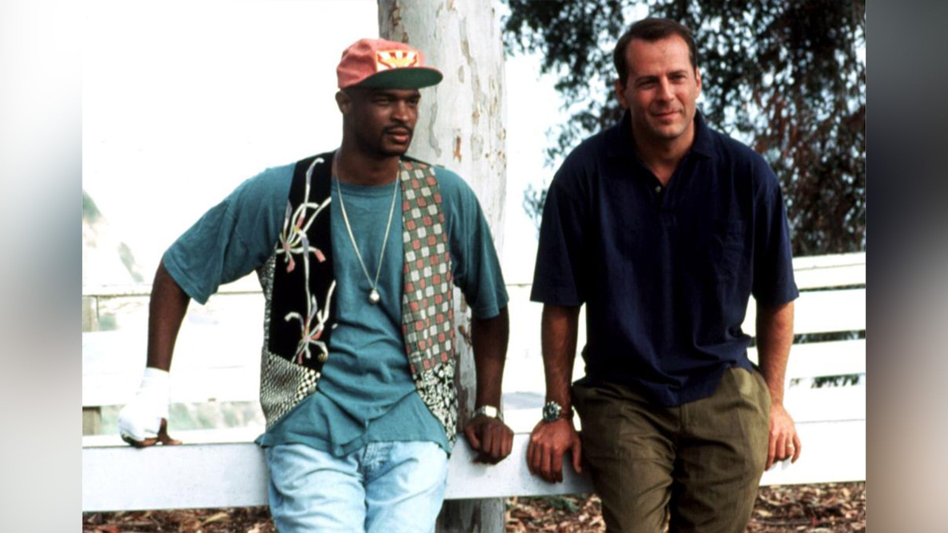 Two men, a dark-skinned and a white man, are leaning on a wooden fence