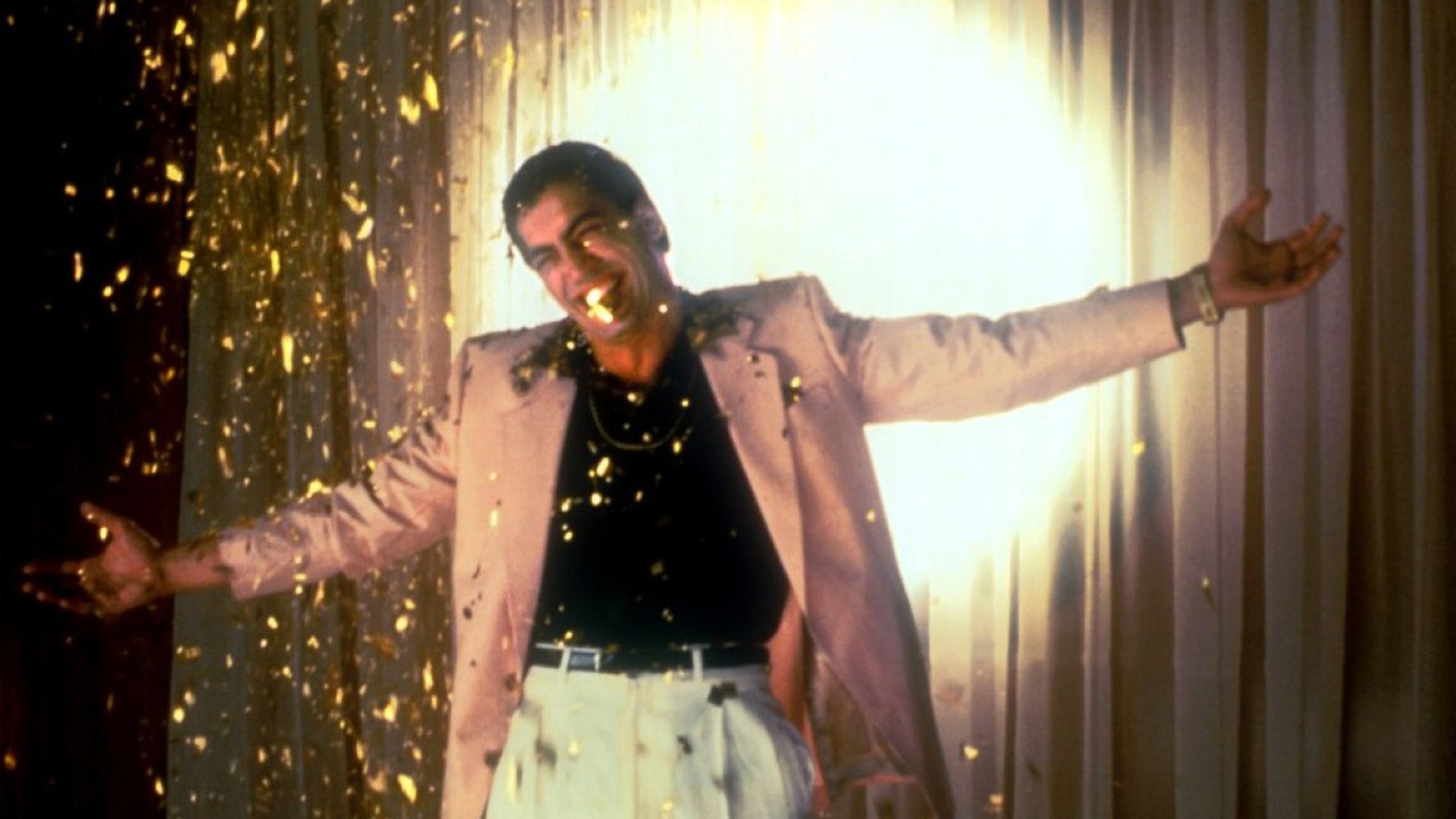 A man dressed in a pink jacket, white trousers, and a black shirt stands with his arms outstretched in a beam of spotlight, with golden confetti in the air