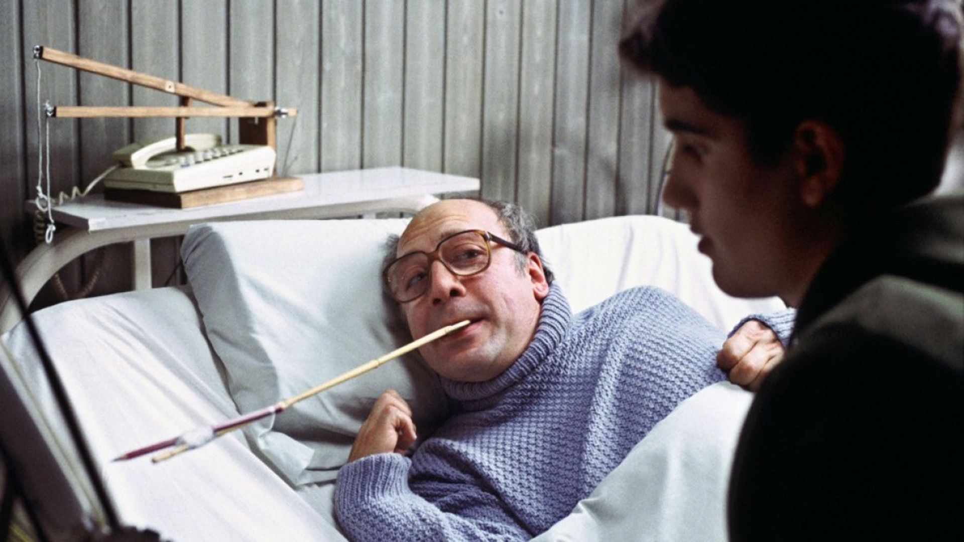 An elderly man in glasses is lying on a hospital bed holding a long stick with a pencil tied to the end in his teeth