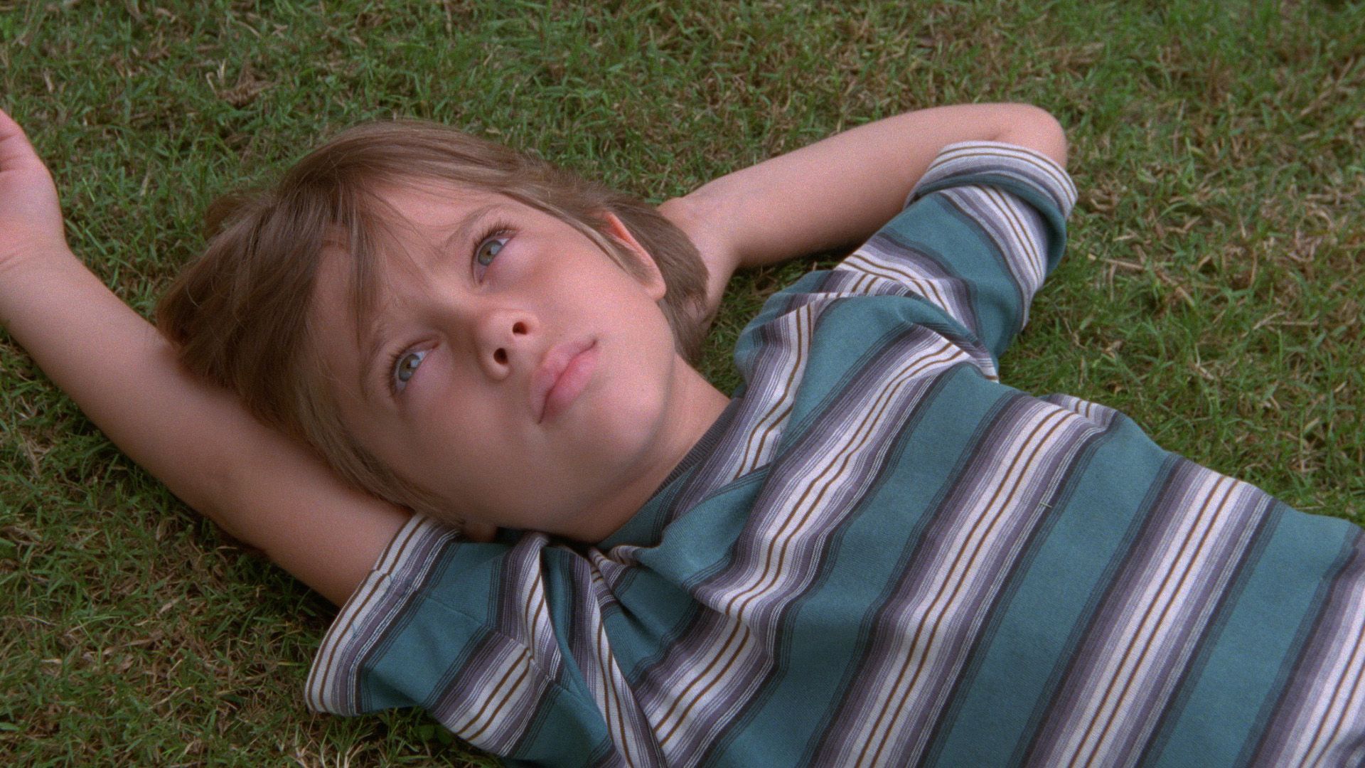 A still from the film "Boyhood": young Ellar Coltrane in a striped T-shirt lying on the grass