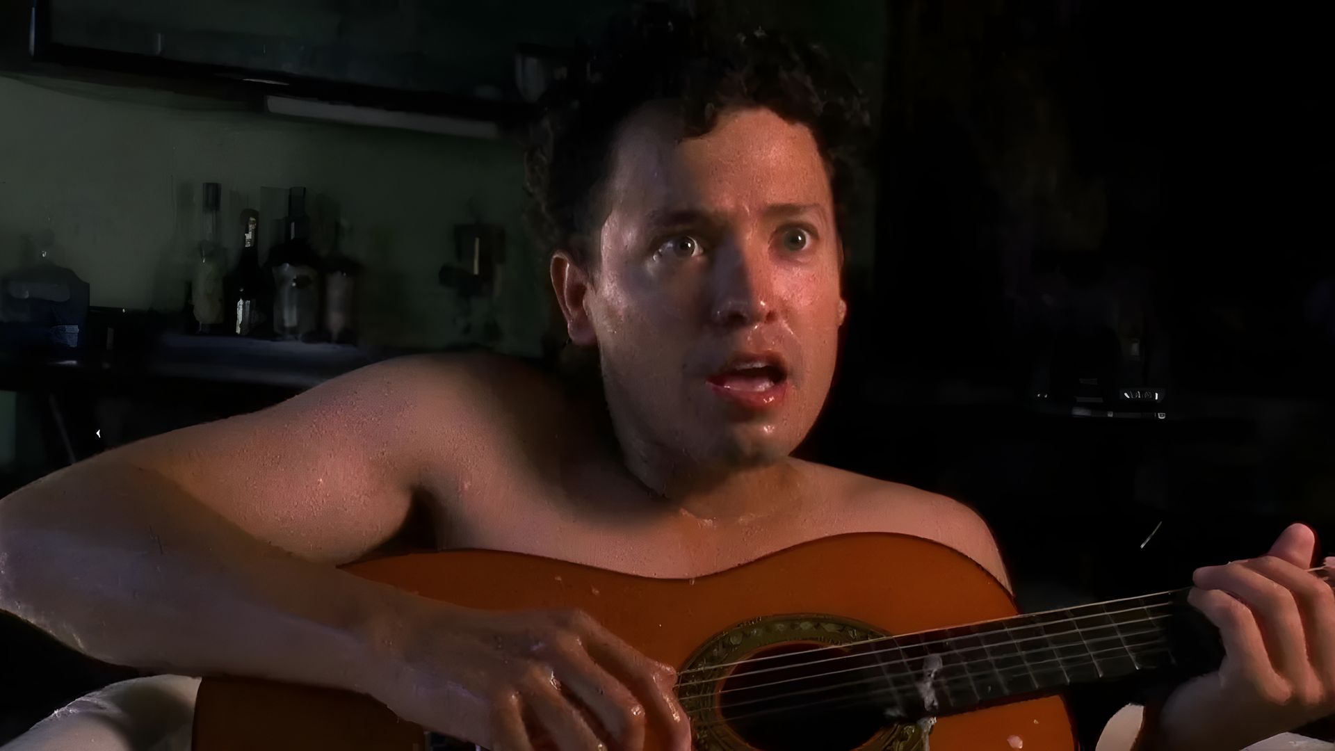 A man without a shirt is playing the guitar, with a surprised expression on his face