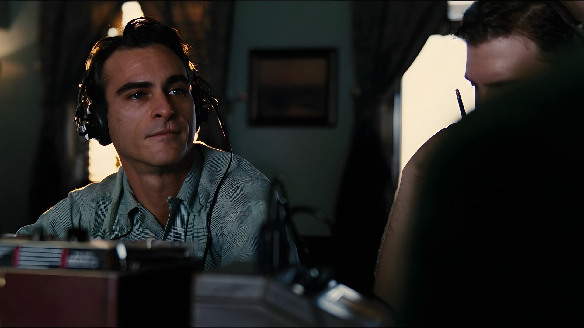 Joaquin Phoenix in a blue shirt and large headphones looks into the distance