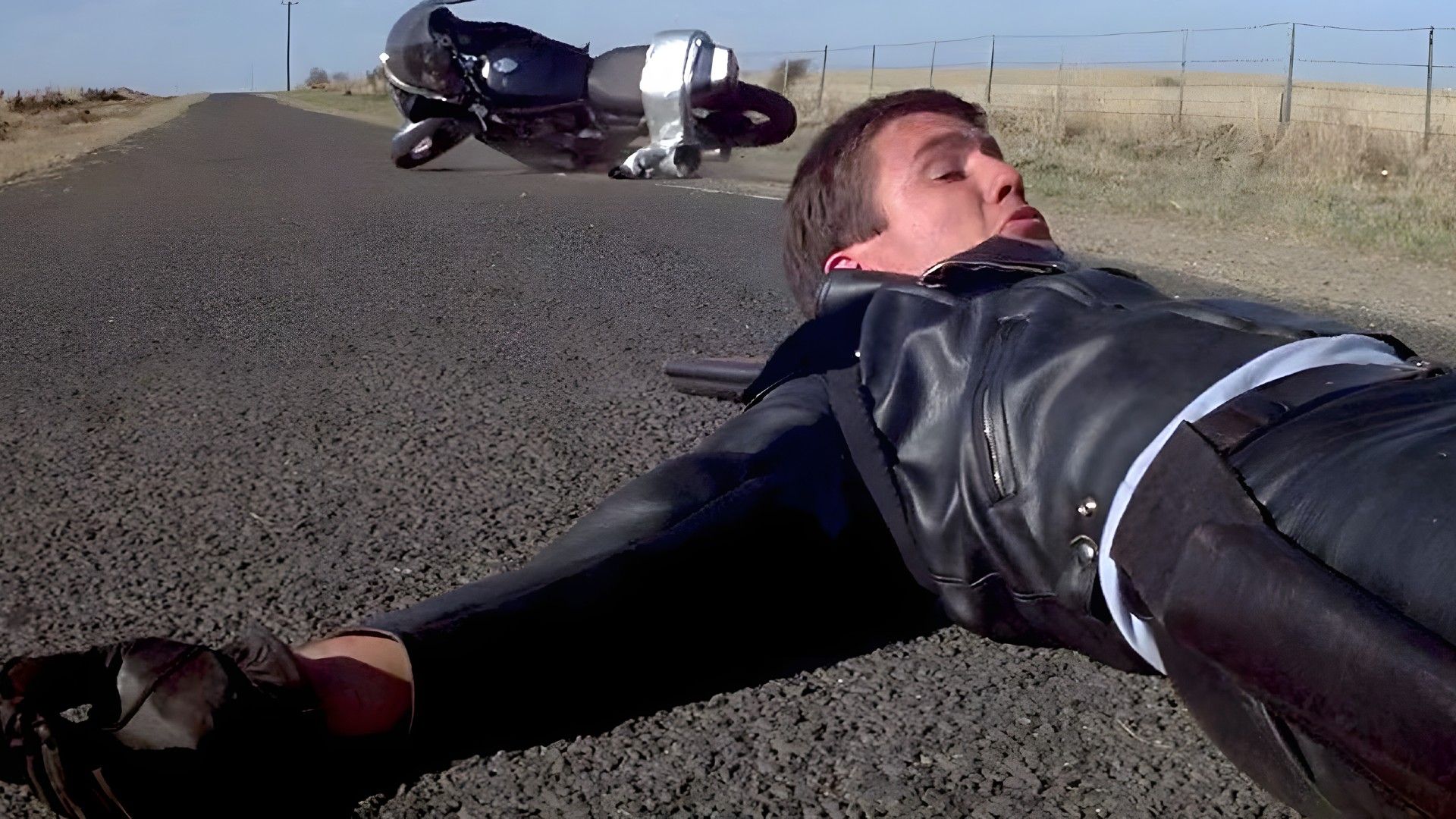 Young Mel Gibson in a motorcycle suit lies on the asphalt, a motorcycle is visible in the background