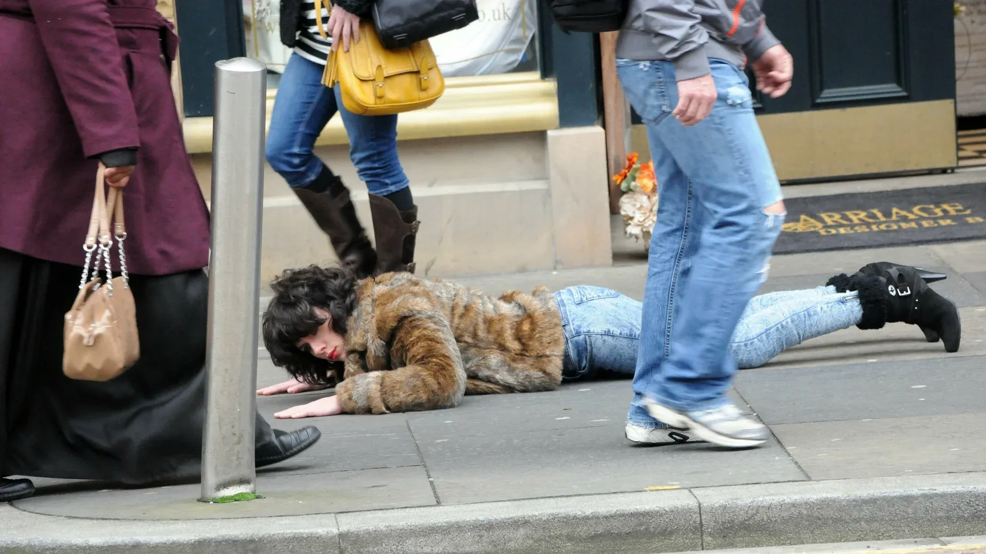 A woman in a fur jacket and jeans is lying on the sidewalk, people walk by without paying attention