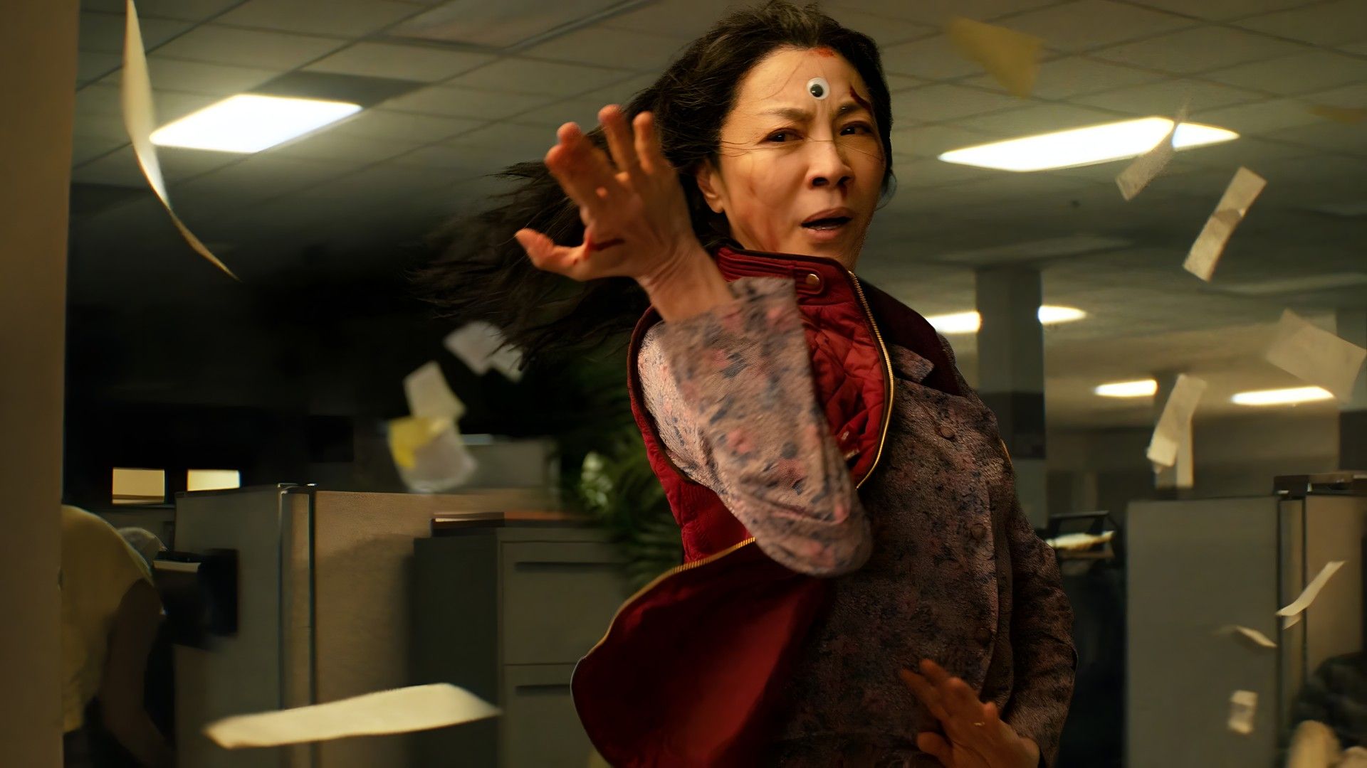 A woman of Asian descent with long black hair in the office, papers flying everywhere. She is wearing a red vest over a colorful blouse and looks focused