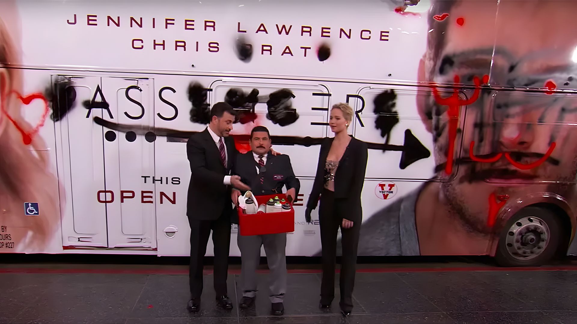 Jimmy Kimmel with an assistant and Jennifer Lawrence are standing by a bus covered in graffiti