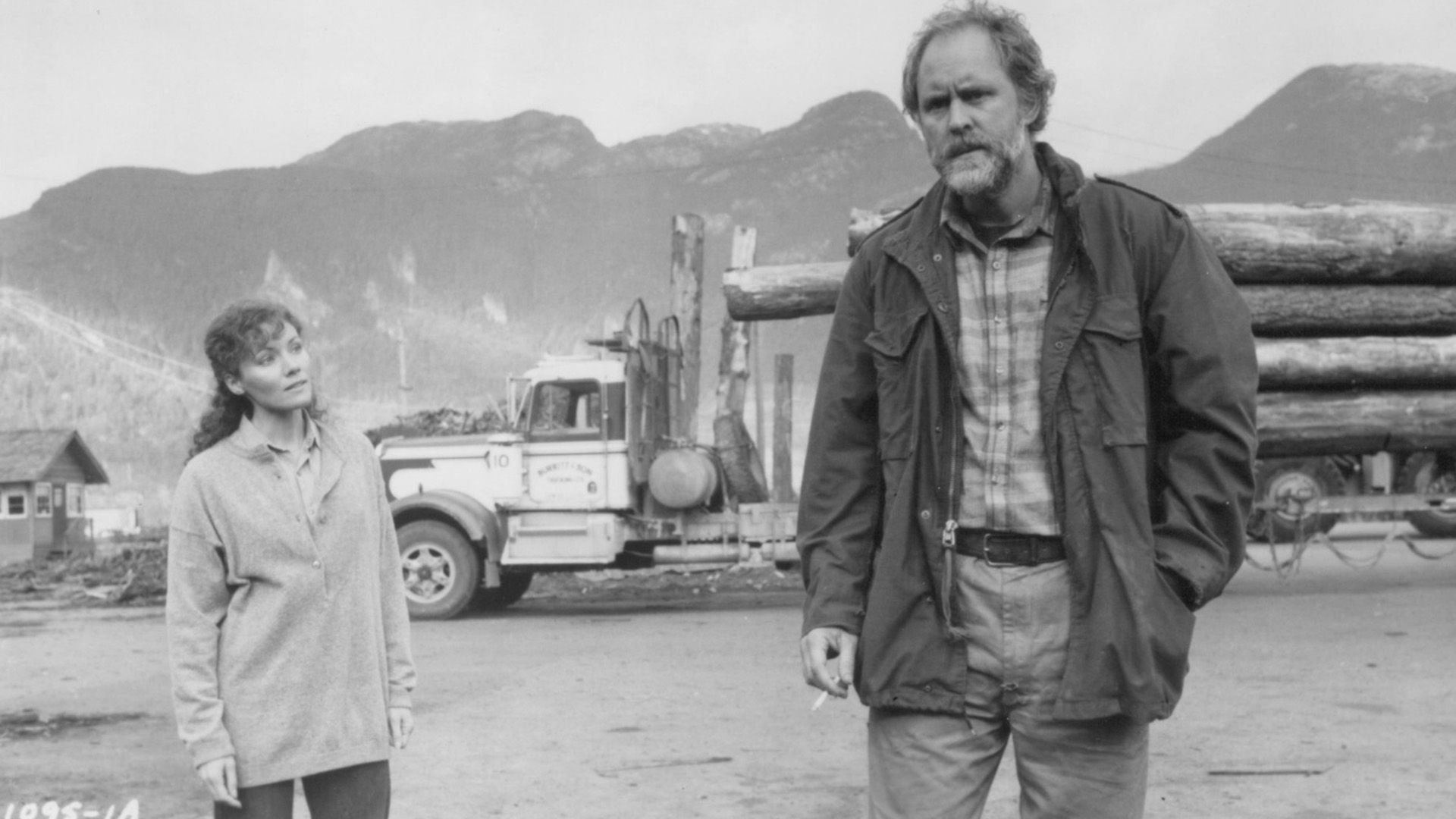 John Lithgow in tattered clothing stands against the backdrop of a logging truck in a scene from the film "Distant Thunder"