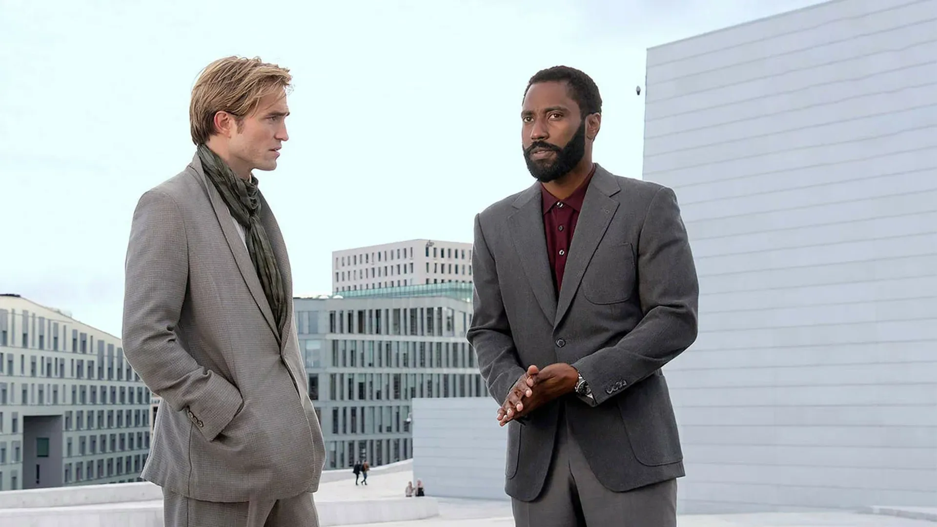 Two men in suits converse against the backdrop of modern buildings. One with a beard gestures with his hand, while the other listens with his hands in his pockets