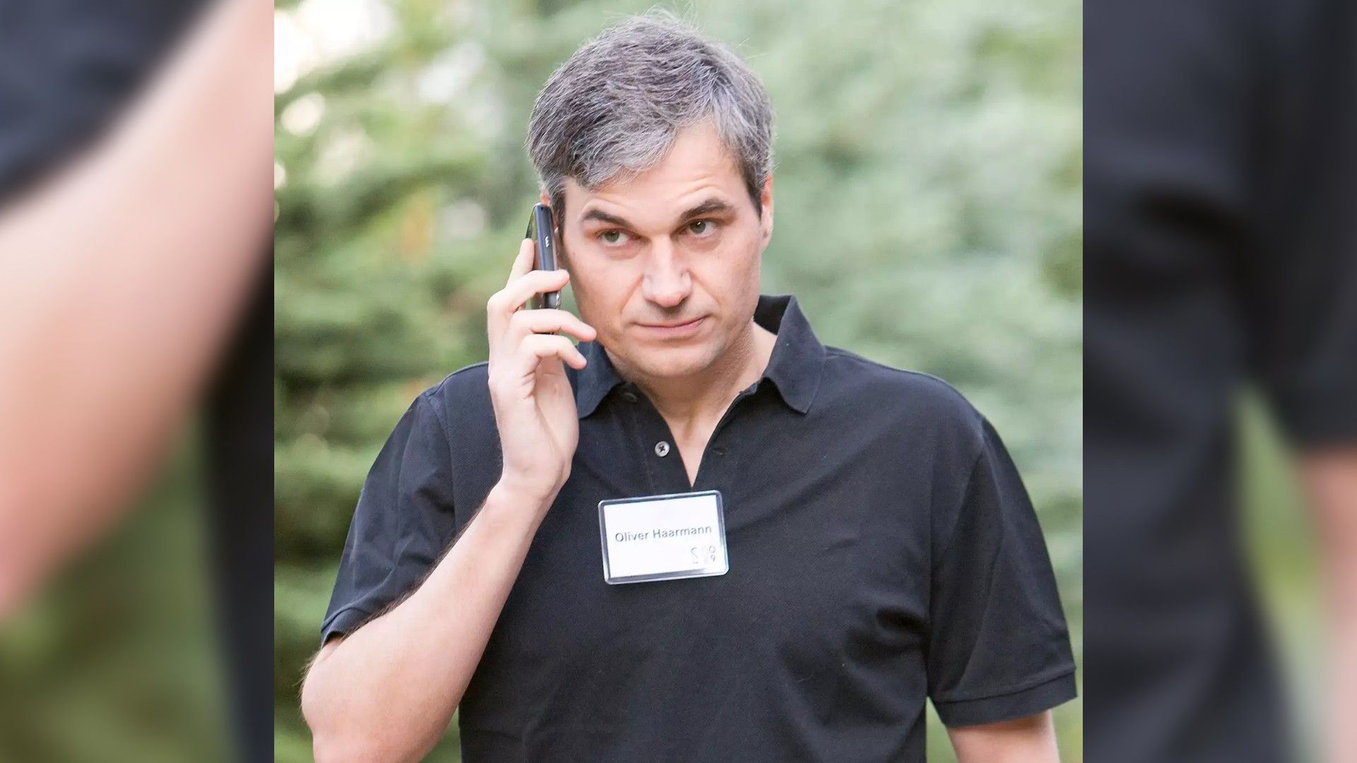 A man in a black shirt with a badge speaking on the phone against a backdrop of green trees, looking focused