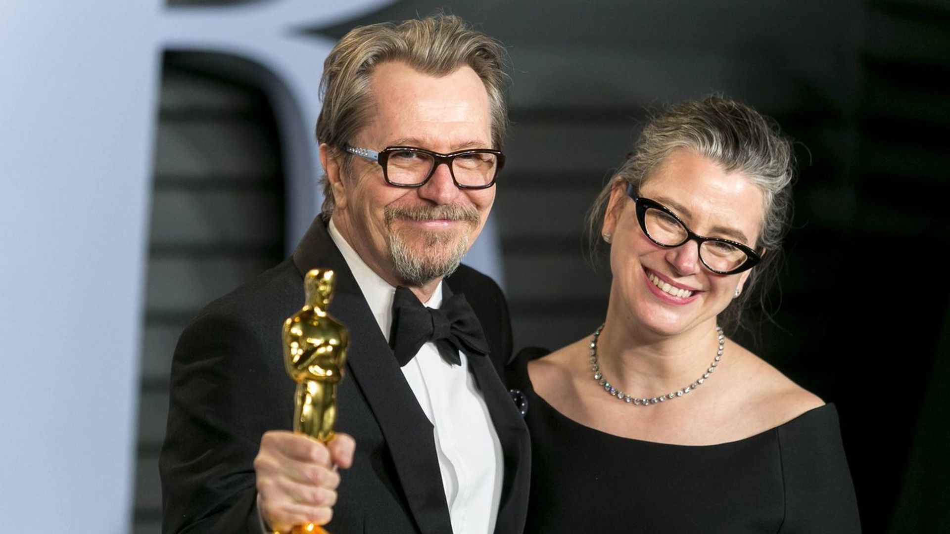 Gary Oldman in a tuxedo holds an Oscar statuette and smiles next to Gisele Schmidt in a black dress and glasses