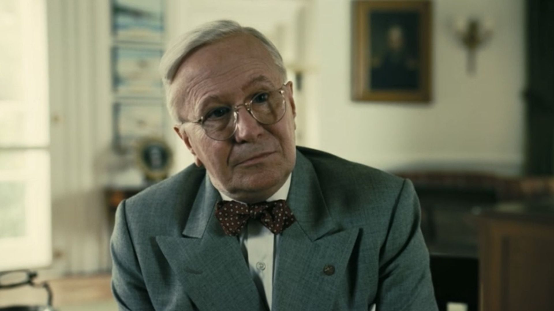 Gary Oldman in glasses and a gray suit with a bow tie sits in an office, looking at the camera with a slight smile