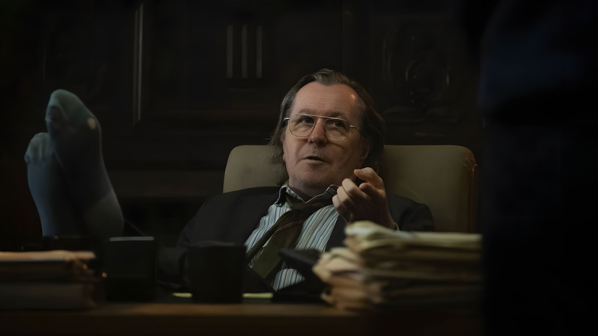 Gary Oldman in glasses and a suit sits at a desk in an office with his feet on the table, wearing a thoughtful expression