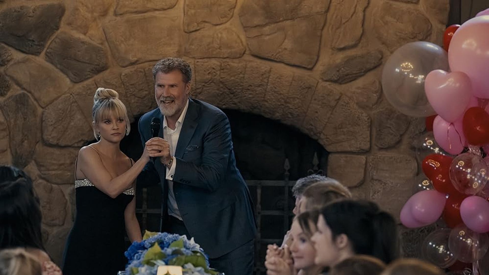 Will Ferrell in a suit with a microphone holds hands with Reese Witherspoon in a black dress against the backdrop of a fireplace, festive table and balloons