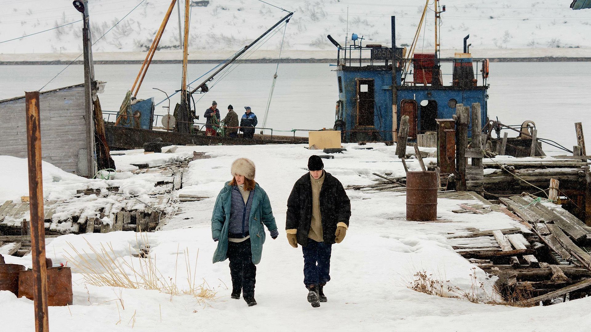 Yura Borisov and Seidi Haarla on a snowy shore against the backdrop of a fishing boat in a scene from the film "Compartment No. 6"