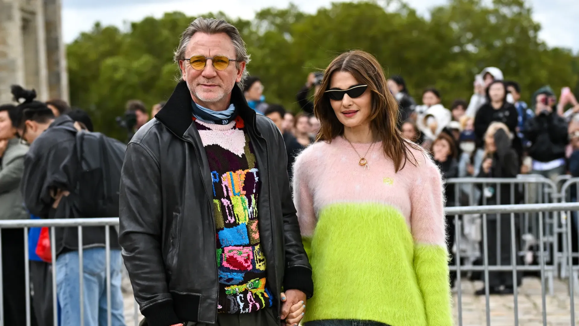 Rachel Weisz in a white and green sweater and Daniel Craig in a black leather jacket and colorful sweater holding hands