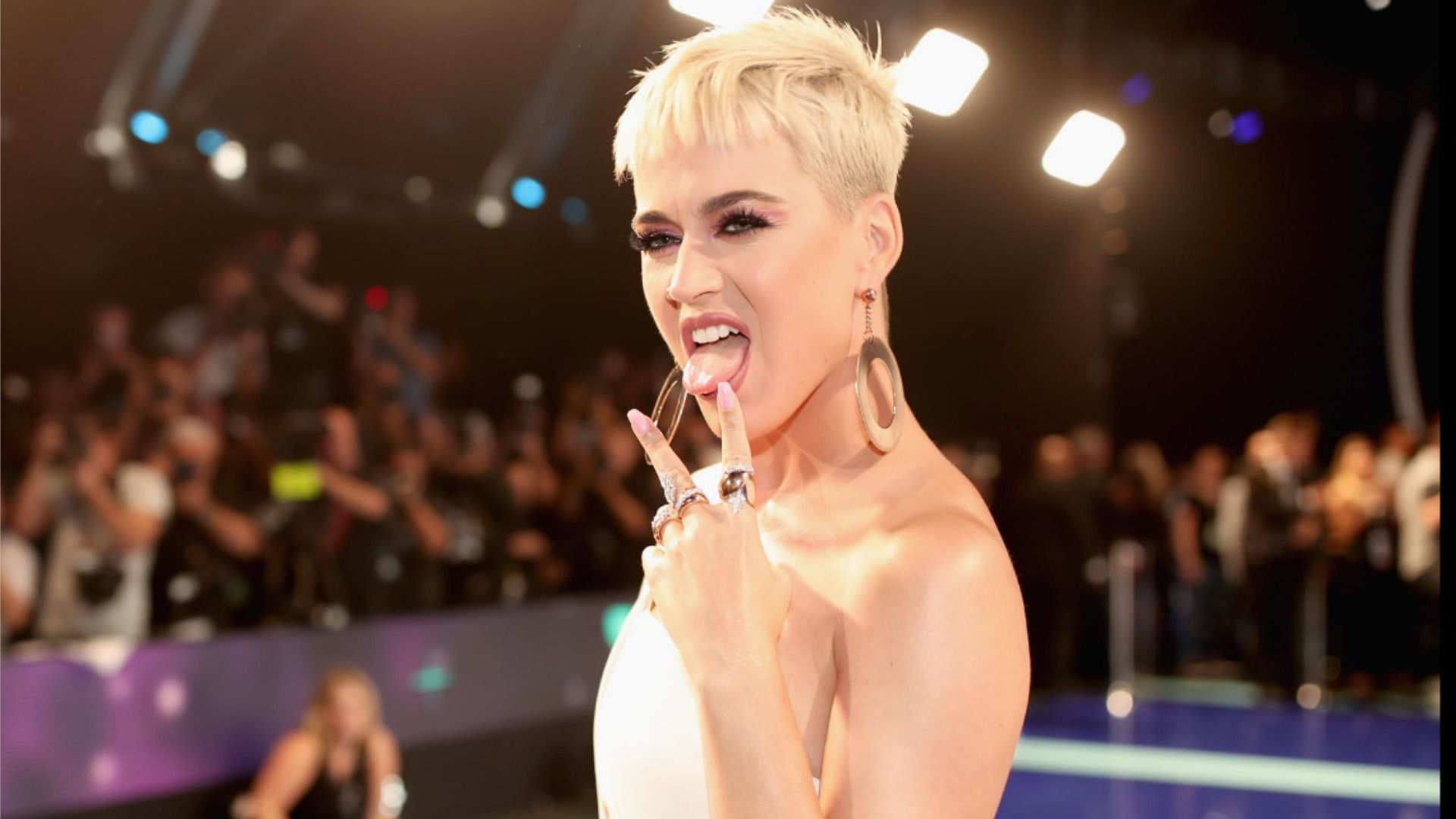 Singer Katy Perry sticks out her tongue and holds up two fingers in a white dress against the backdrop of the audience