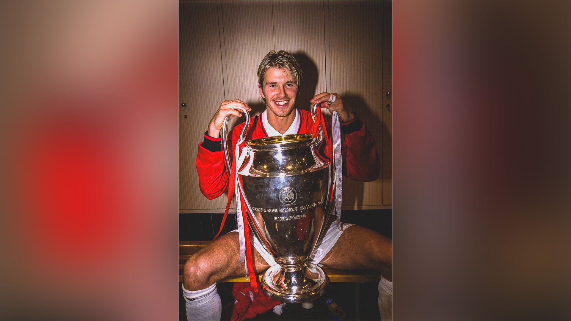Young David Beckham sitting on the bench of the sports locker room with the UEFA Cup