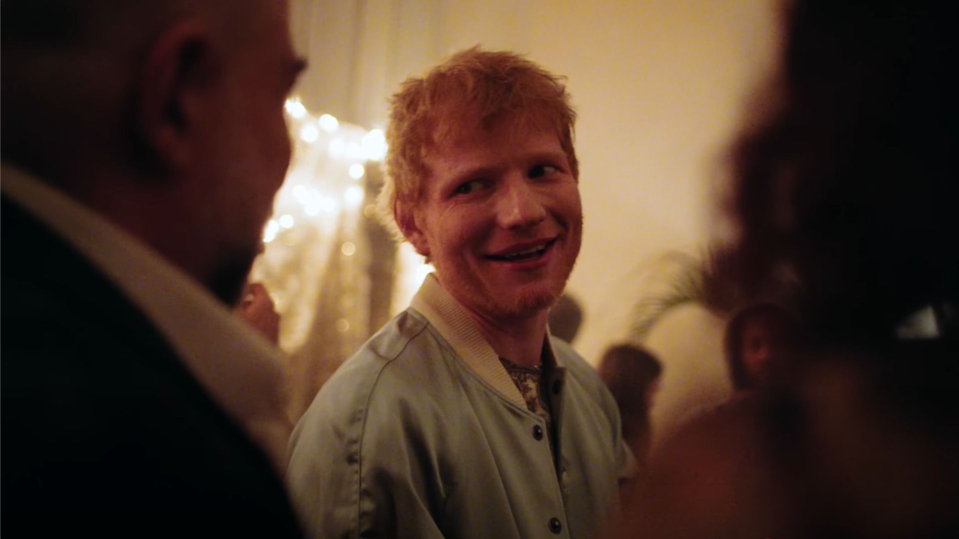 Singer Ed Sheeran looks and smiles in the yellow-orange light next to the figures of people