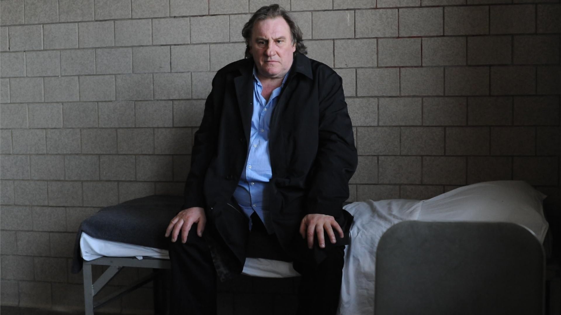 Actor Gérard Depardieu in a classic suit sits on a small bed and looks at the floor