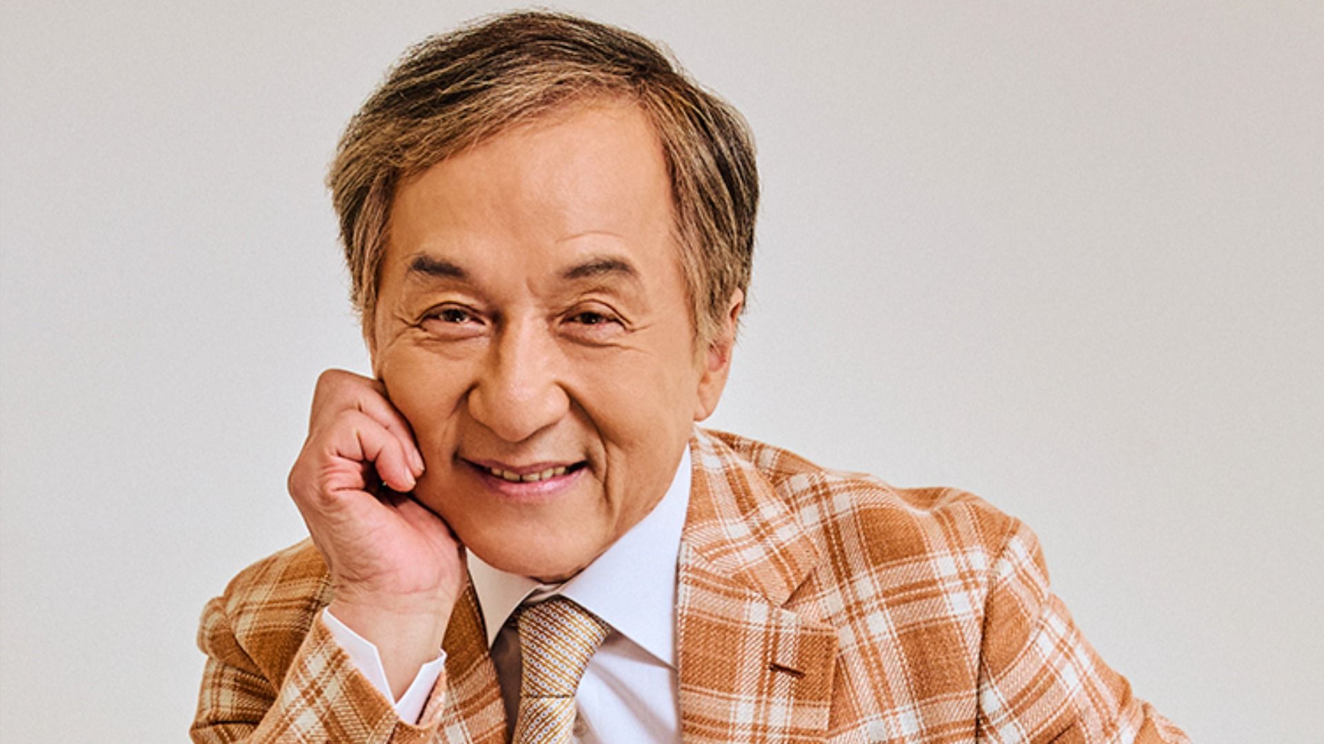 Actor Jackie Chan in a checkered brown jacket sits, leaning on his hand, and smiles at the camera against a white background