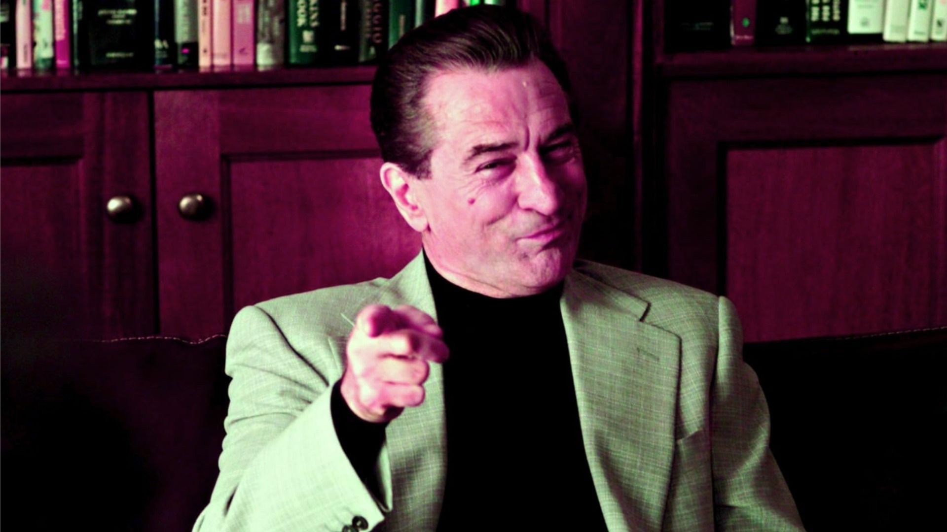Actor Robert De Niro in a gray jacket is pointing with his index finger and smiling against the backdrop of a bookshelf