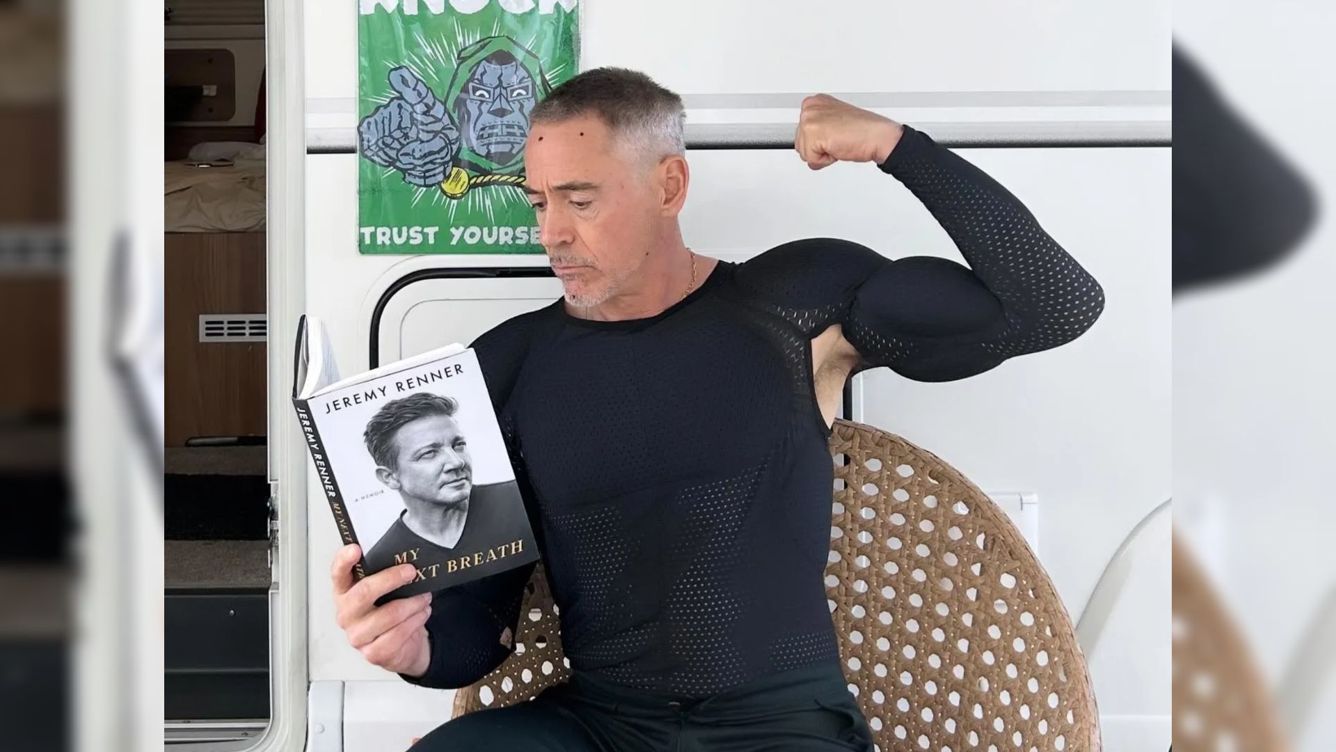 Actor Robert Downey Jr. in tight clothing sits by a trailer and holds a book in his hands