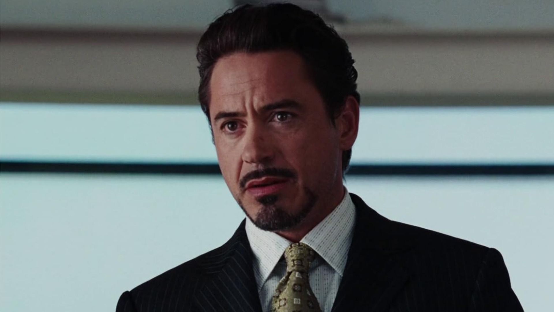 Actor Robert Downey Jr. with a mustache and beard stands in a classic suit and looks to the side against the background of a window