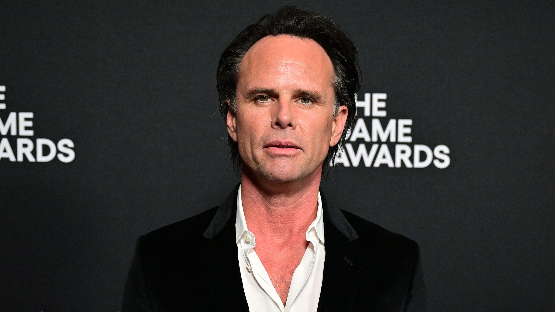 Actor Walton Goggins in a black jacket and white shirt with an unbuttoned collar against a black background
