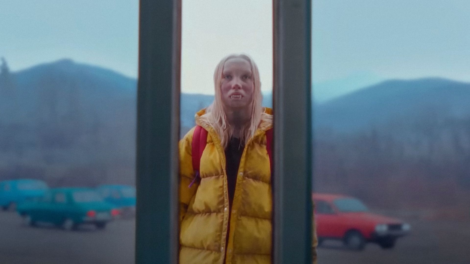 A girl in a yellow jacket with vampire teeth stands at the supermarket doors