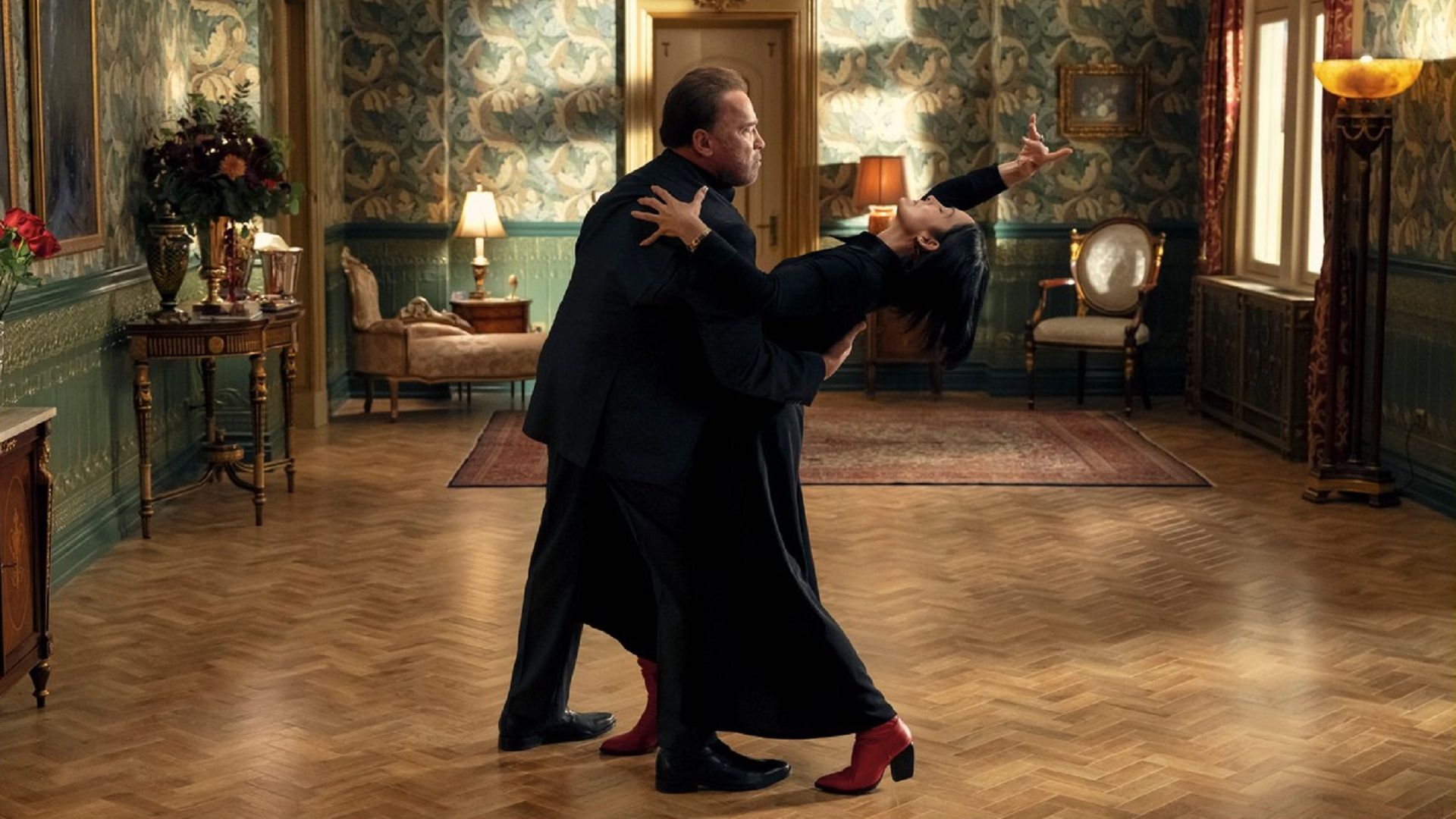 Arnold Schwarzenegger and Carrie-Anne Moss dancing in black suits in the middle of the room