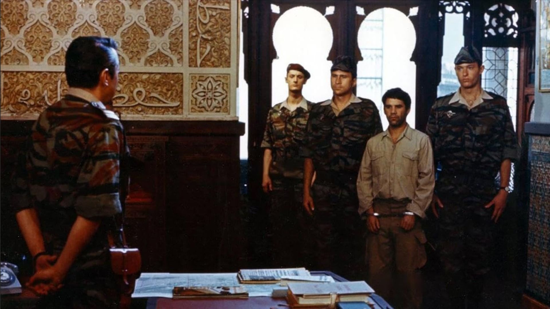 A man with handcuffs on his hands stands surrounded by soldiers at a table