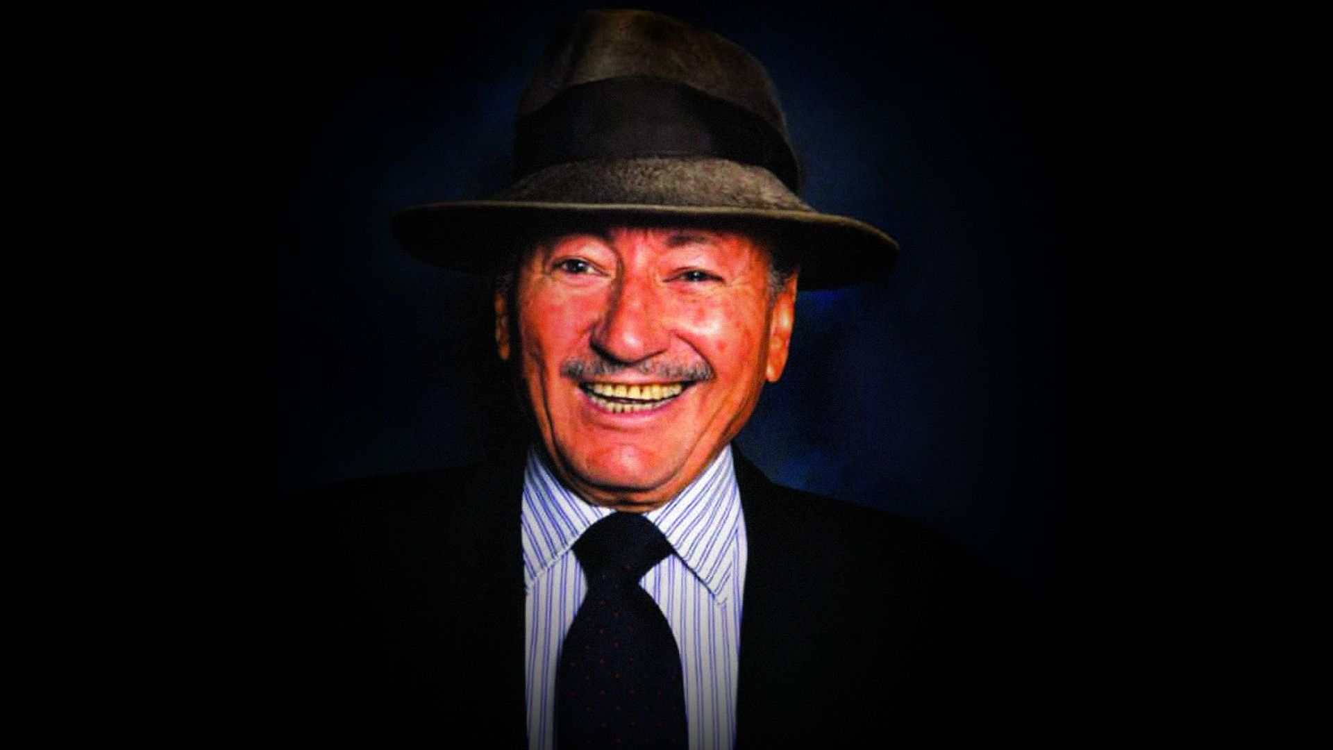 A smiling man in a hat and jacket looks at the camera against a black background