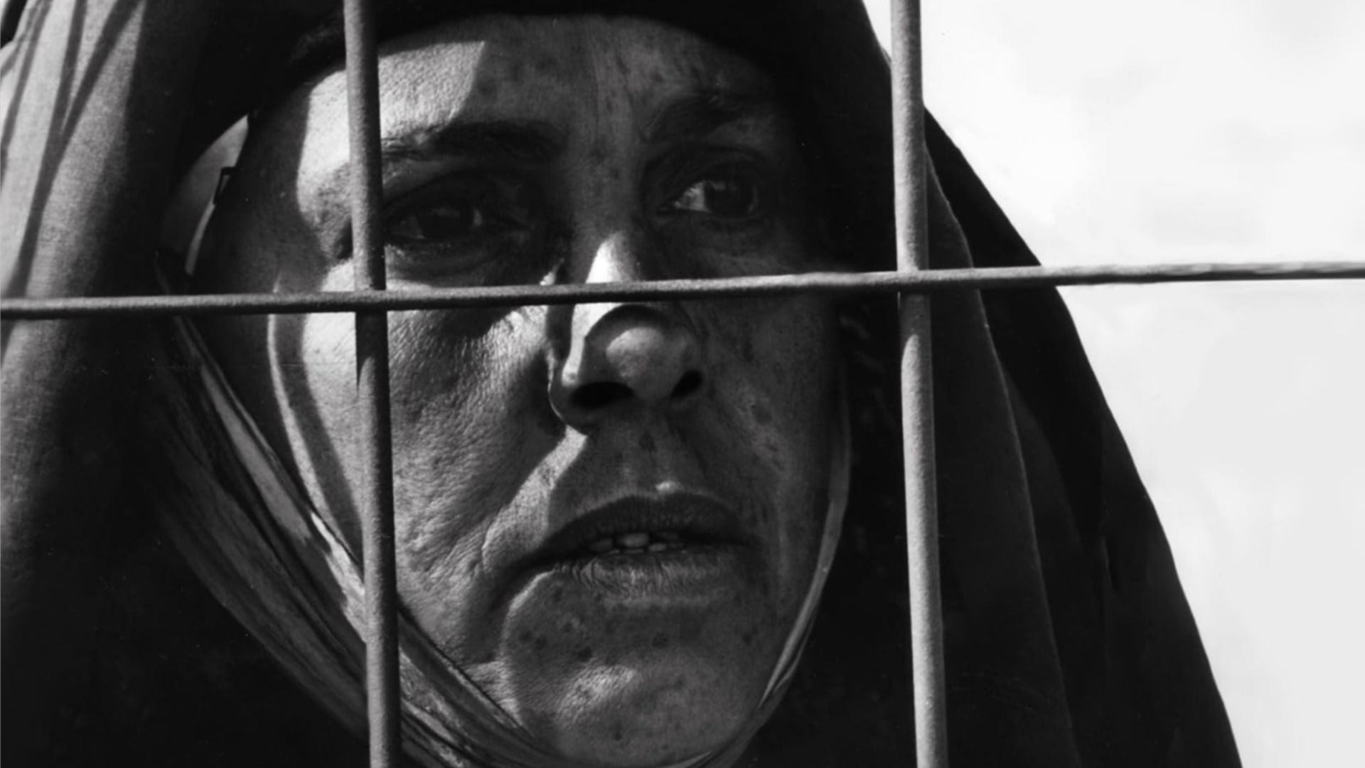 A woman with her head covered stands behind an iron mesh and looks in the direction