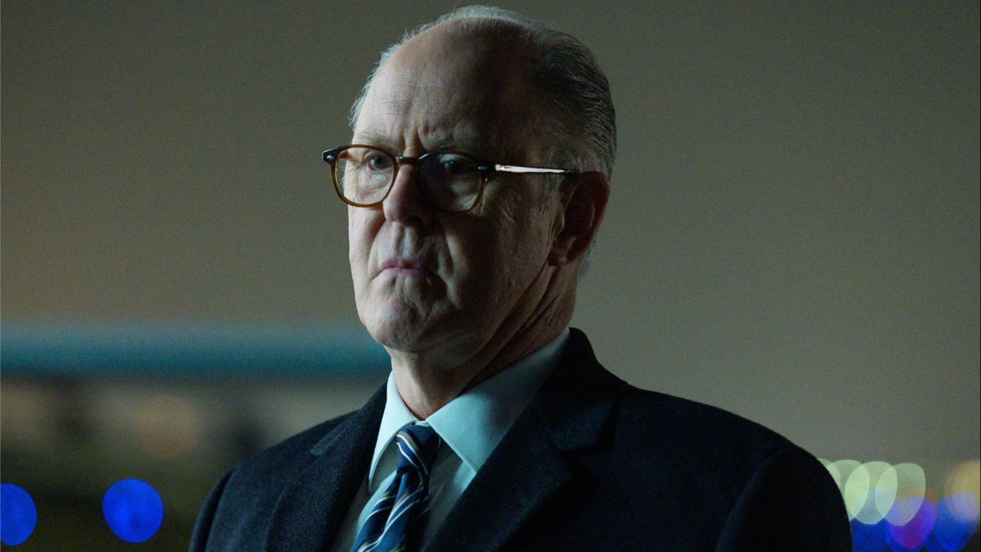 Actor John Lithgow in a suit and glasses stands emotionless and looks to the side