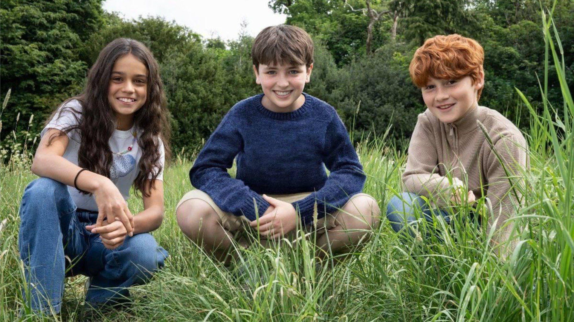 Actors Dominic McLaughlin, Alastair Stout, and Arabella Stanton sitting on the grass and smiling