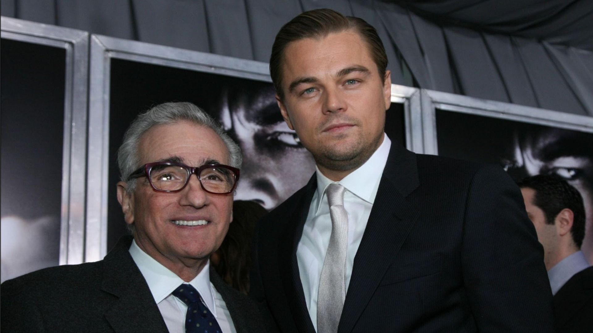 Director Martin Scorsese wearing glasses stands and smiles next to actor Leonardo DiCaprio, both in black suits against a backdrop of posters