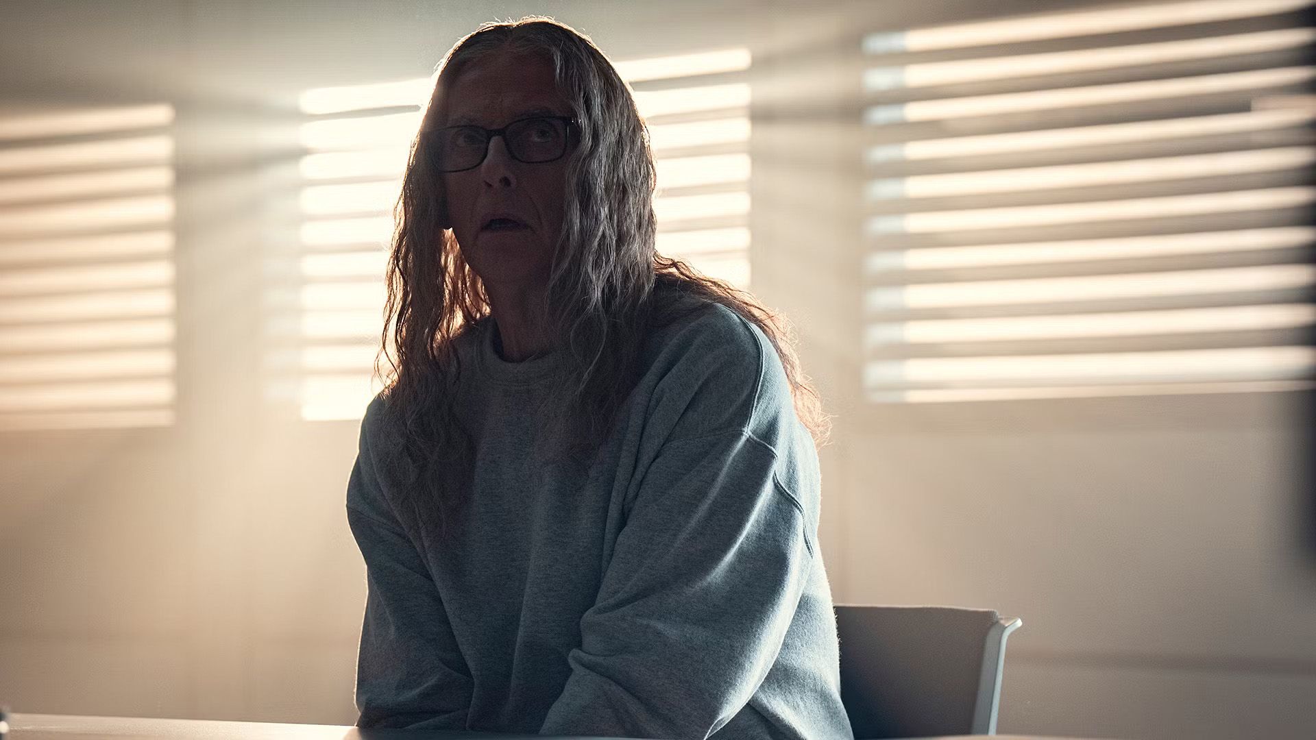Peter Capaldi with long gray hair and glasses sits at a table against a backdrop of windows with pouring light, looking to the side