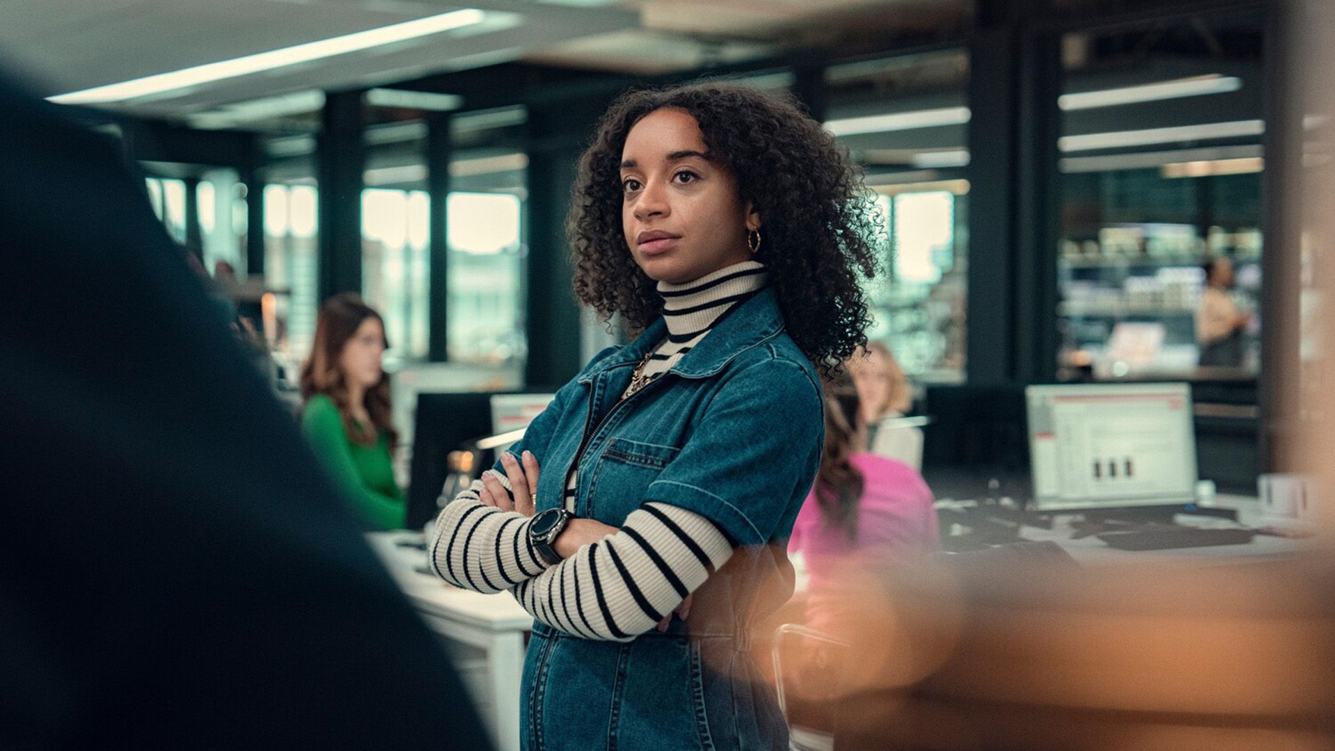 Siena Kelly in a striped sweater and denim shirt stands in the middle of the office, arms crossed over her chest
