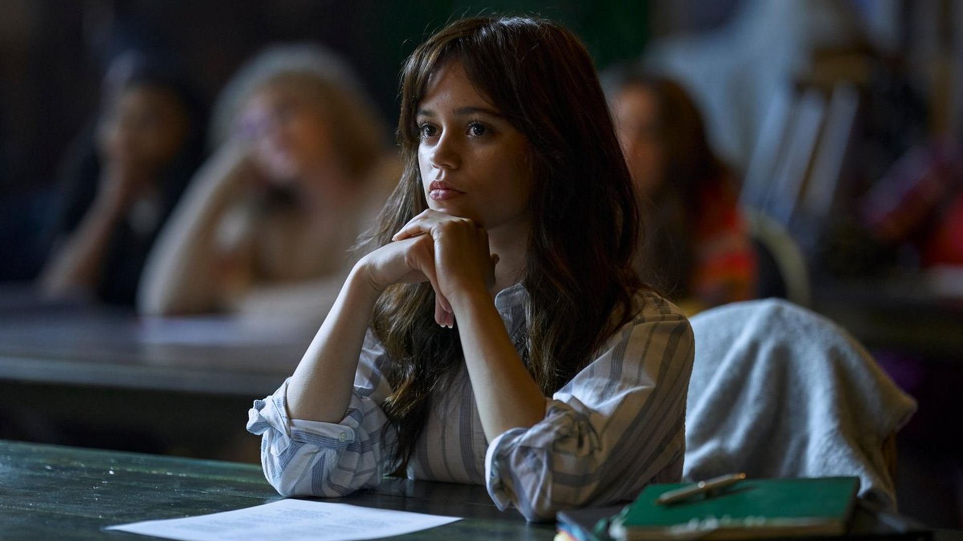 Jenna Ortega in a white sweater sits at a desk during class with a focused expression