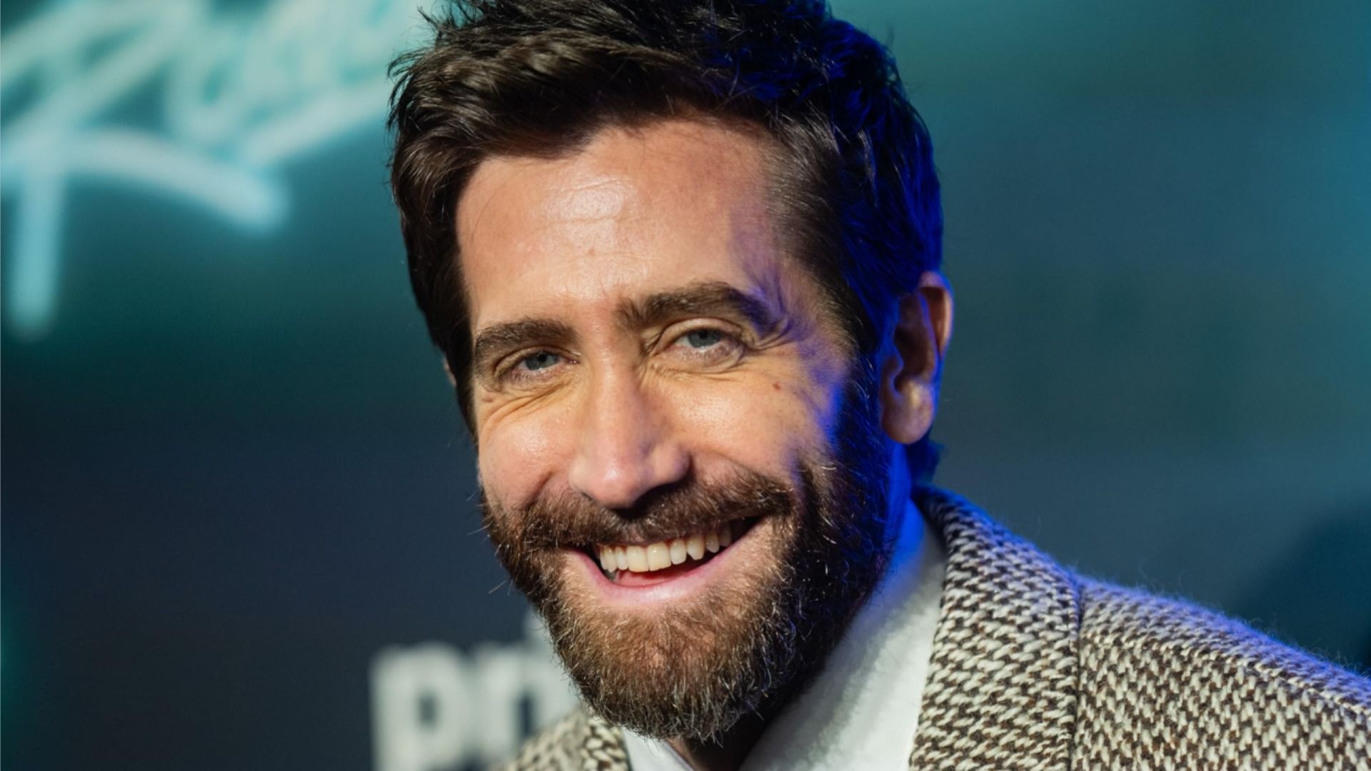 Actor Jake Gyllenhaal in a gray blazer smiles against a blue background