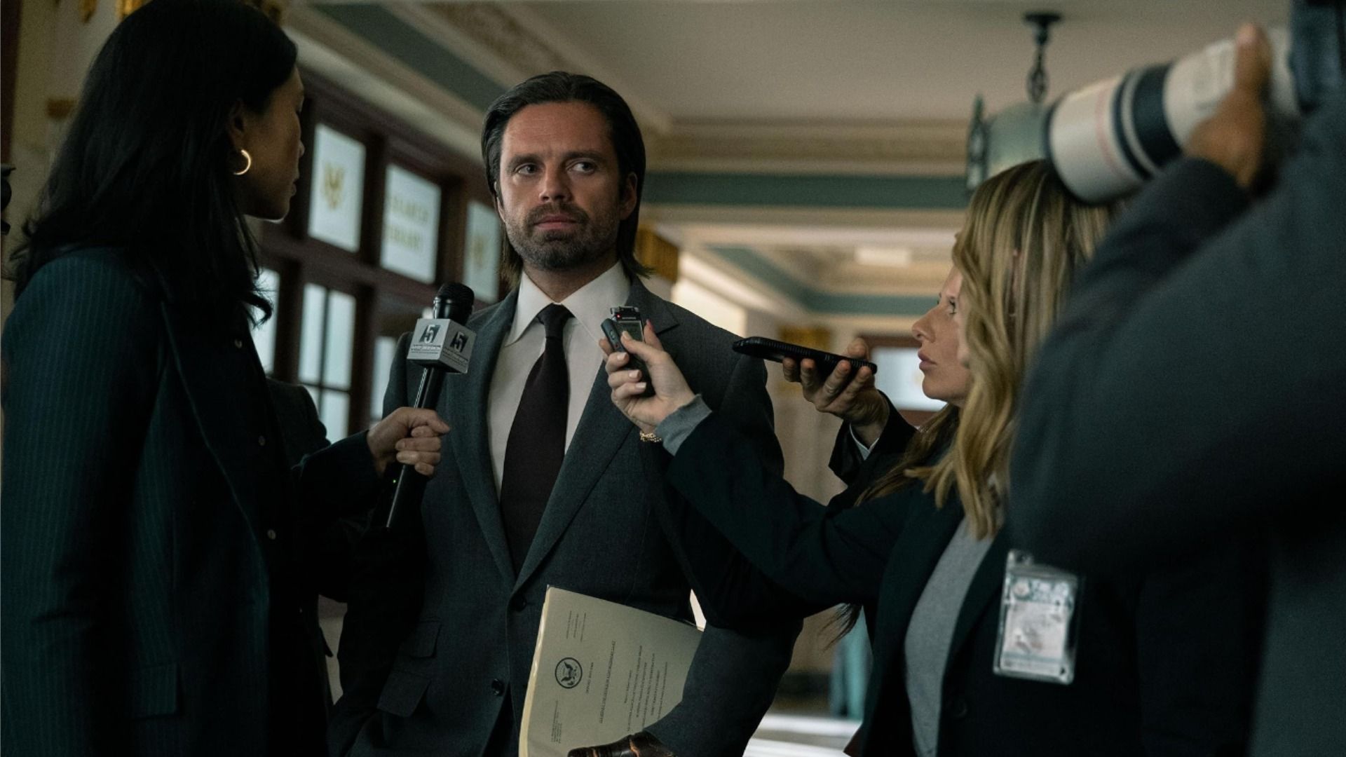 Actor Sebastian Stan in a black suit with long hair surrounded by journalists